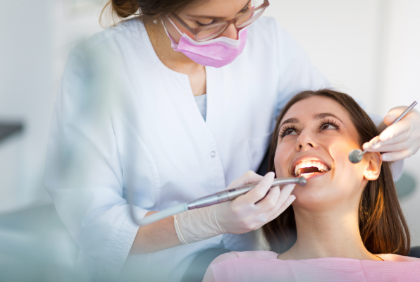 A female dentist in a white coat and green mask performing a visual oral cancer screening on a young female patient in a dental chair.