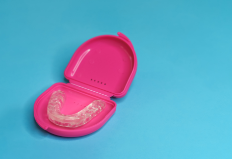 A clear plastic orthodontic retainer sitting inside an open bright pink protective plastic storage case on a blue background.